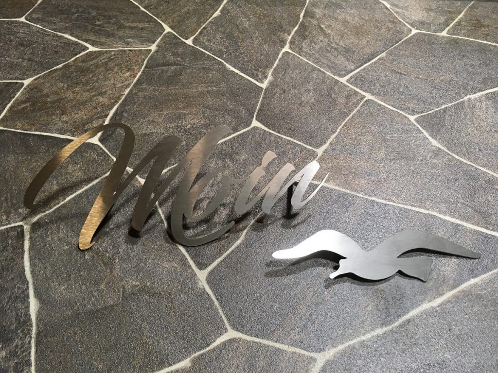 Metall-Schriftzug Moin mit ausgeschnittener Möwe aus Edelstahl auf Natursteinboden
